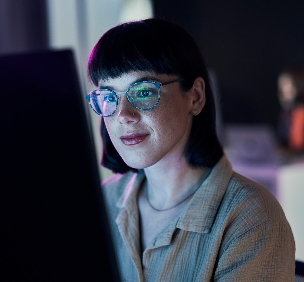 Business woman looking at data on a computer at night. The coding is reflecting off of her glasses.