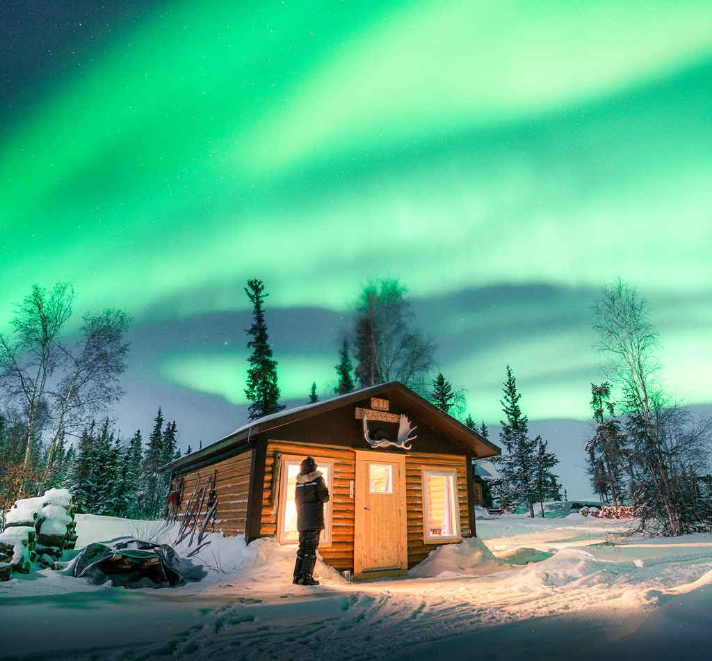 A traveller standing outside a cabin looking at the northern lights in Yellowknife, Northwest Territories, Canada.